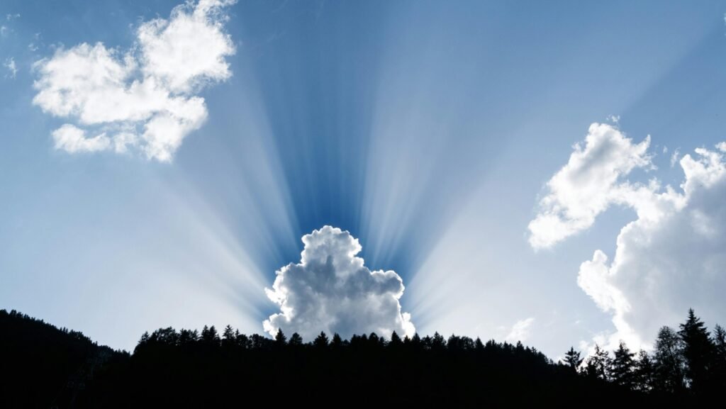 A stunning natural phenomenon as sunrays pierce through fluffy clouds above a silhouetted forest.