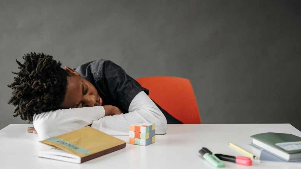 A young boy sleeping at a desk with books and a Rubik's cube, looking weary.