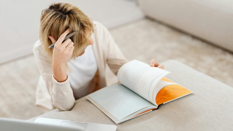 Blonde woman studying in living room with a book and laptop, creating a relaxed atmosphere.